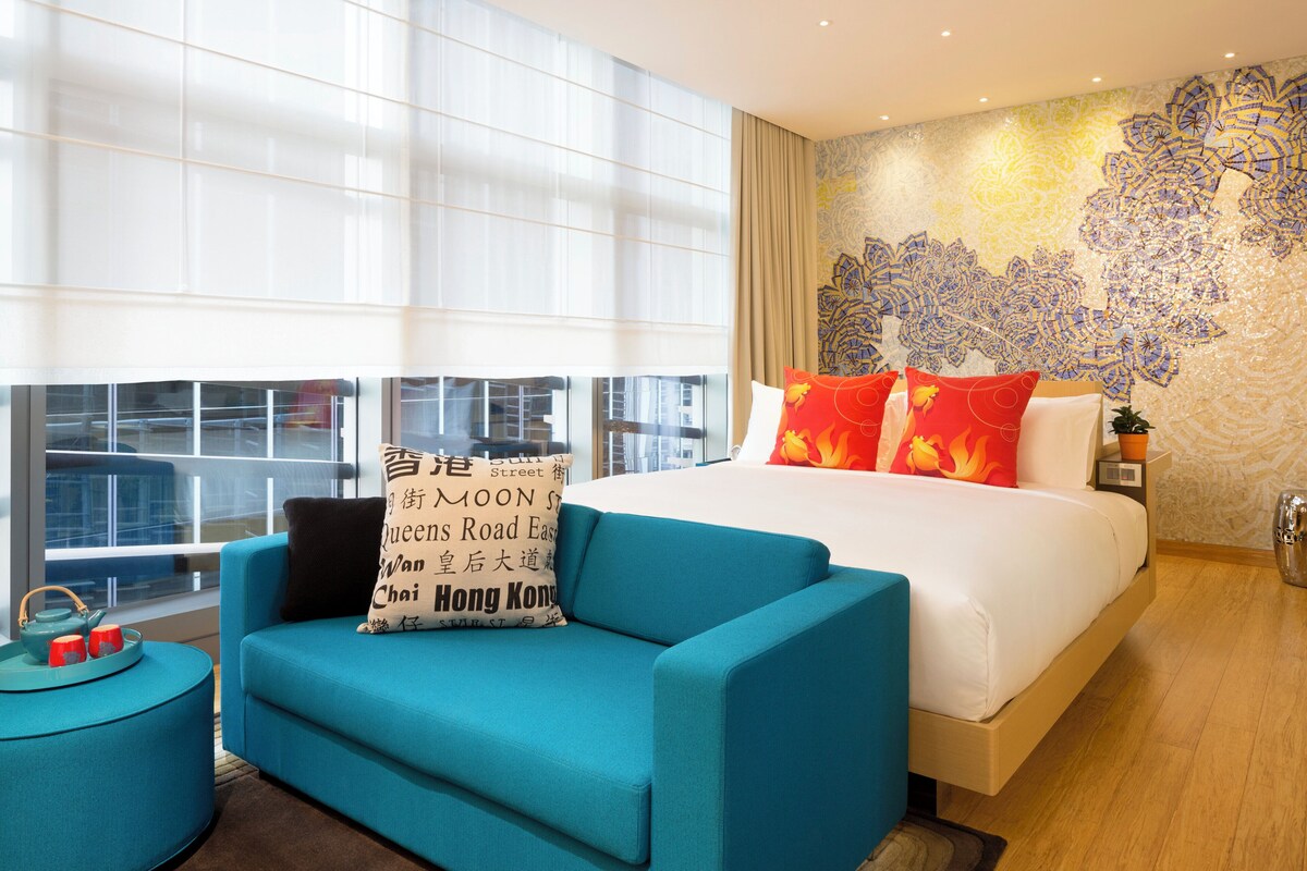 Un hotel moderno con una cama grande, un sofá azul, una mesita con tetera y vistas a la ciudad a través de amplios ventanales.