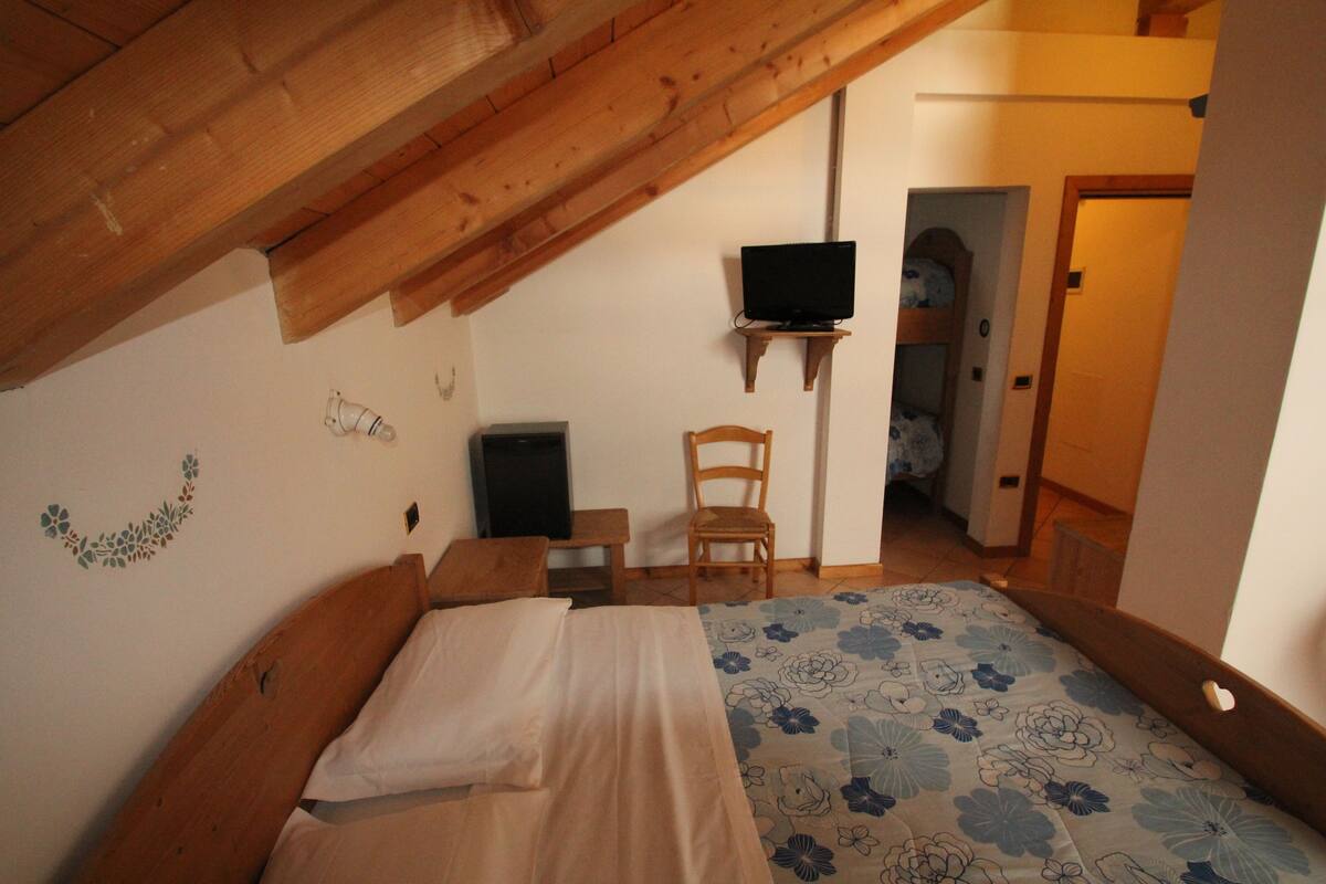 A bedroom with a wooden ceiling, a bed with a floral bedspread, a television on a stand, and a chair.