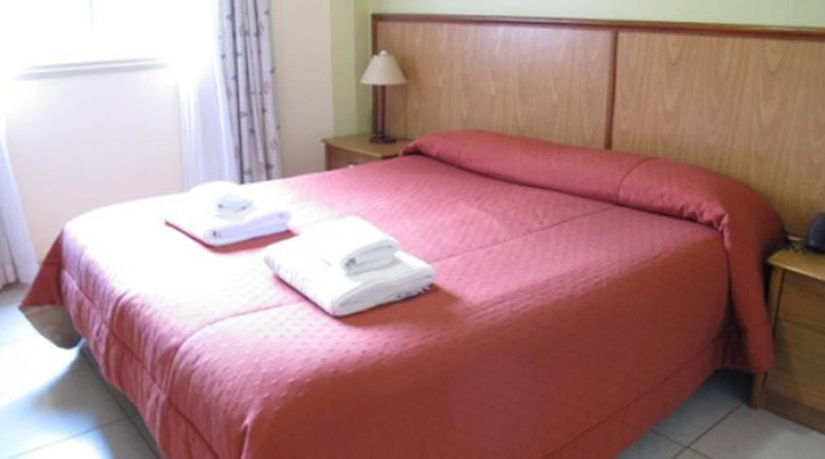 A neatly made bed with a red bedspread, a white pillow, and a folded white towel on the bed.