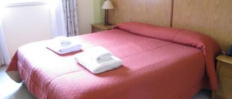A neatly made bed with a red bedspread, a white pillow, and a folded white towel on the bed.