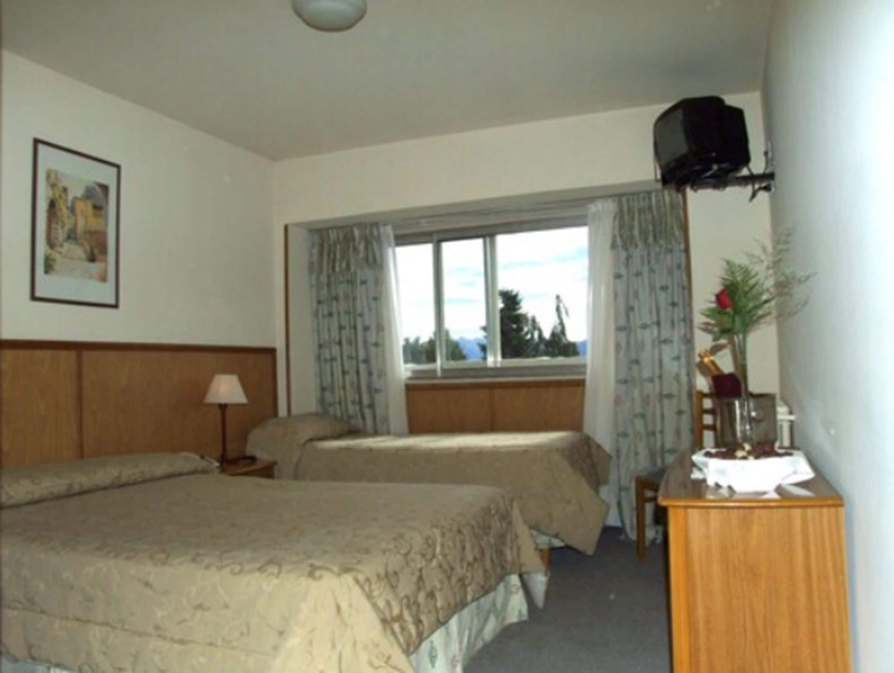 A hotel room with two beds, a window with curtains, a lamp, a television on the wall, and a painting on the wall.