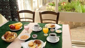 A breakfast table with various food items and drinks, set outdoors with chairs and a view of a garden.