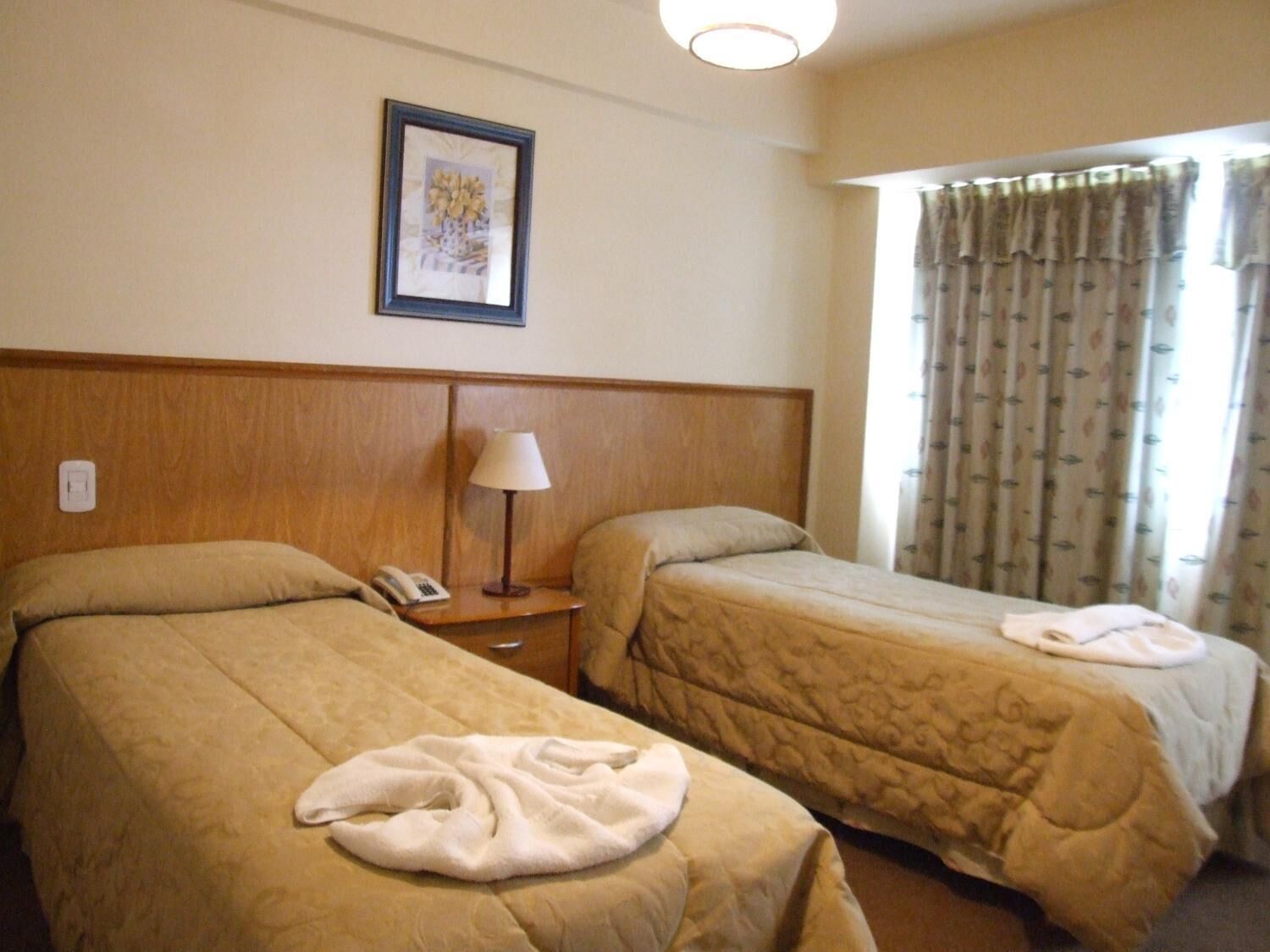 Une chambre d’hôtel avec deux lits, une table de chevet avec une lampe, un tableau encadré au mur et une fenêtre avec des rideaux.
