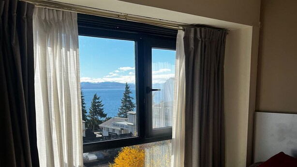 A hotel room with a bed, a window with curtains, and a view of the sea and mountains.