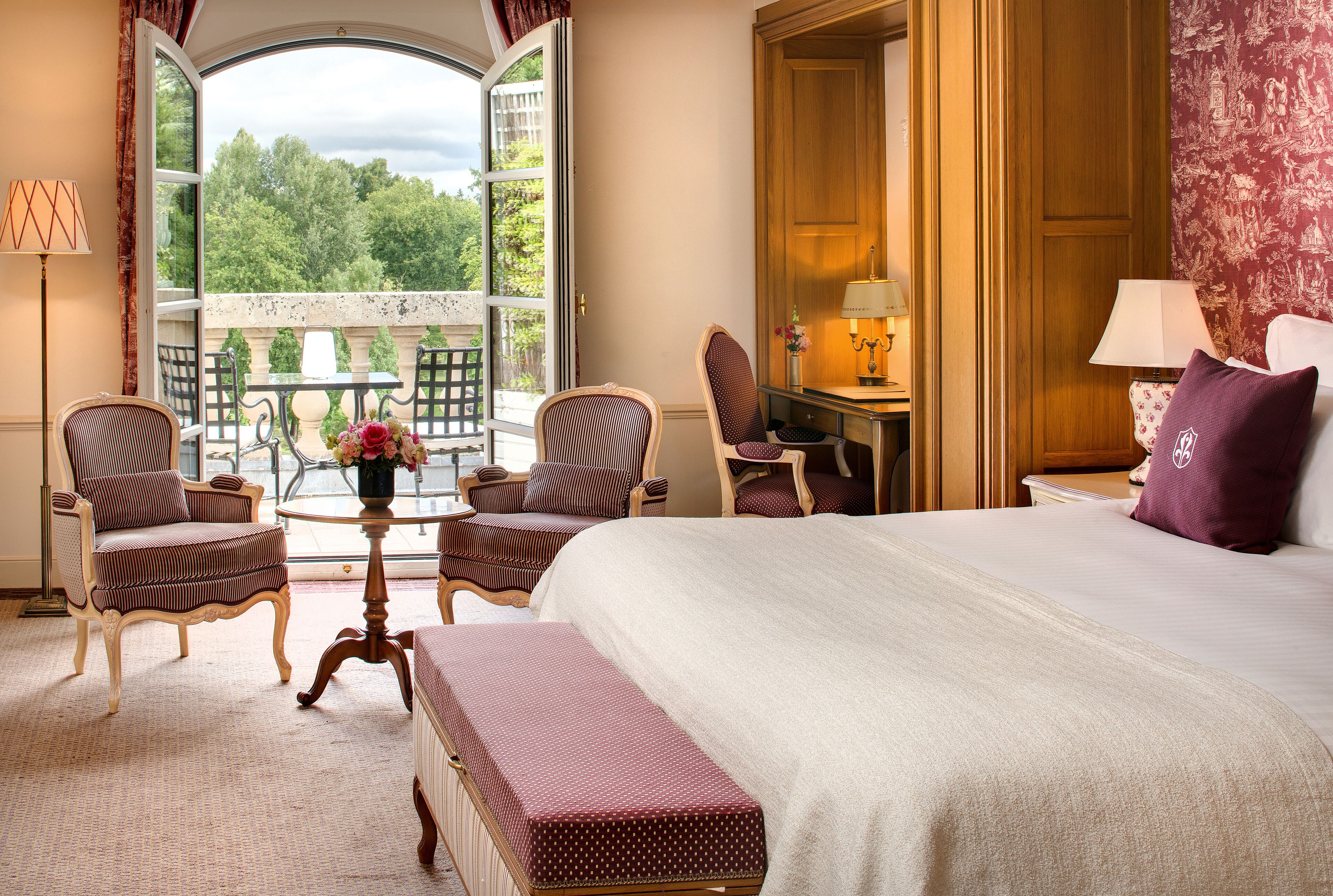 Une chambre d’hôtel avec un grand lit, deux fauteuils, une petite table avec un vase, un bureau avec une lampe et une vue sur la verdure par une fenêtre ouverte.