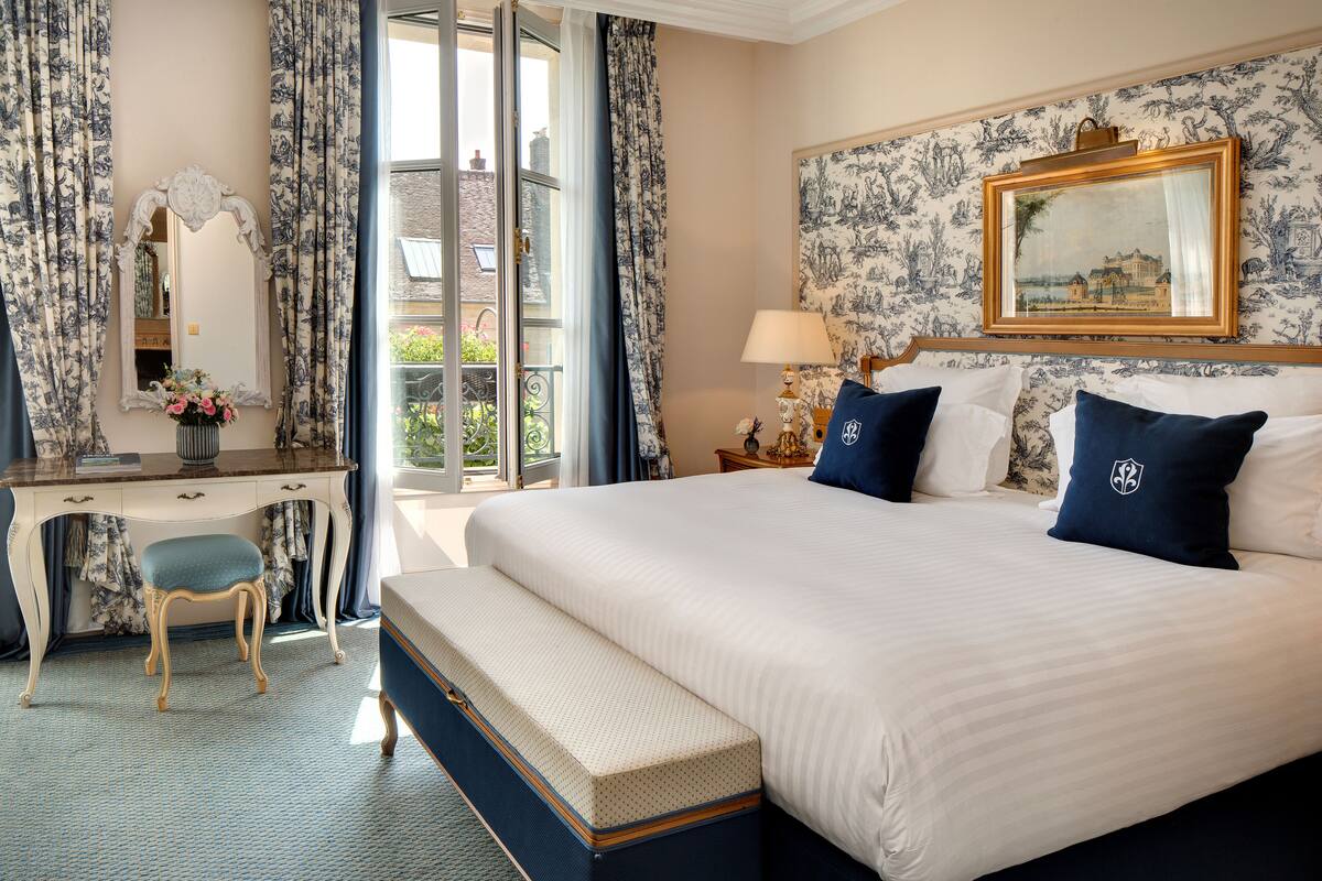 Ein Schlafzimmer mit einem großen Bett, einem Schreibtisch, einem Sessel, einer Bank, einem Spiegel und einem Fenster mit Vorhängen.