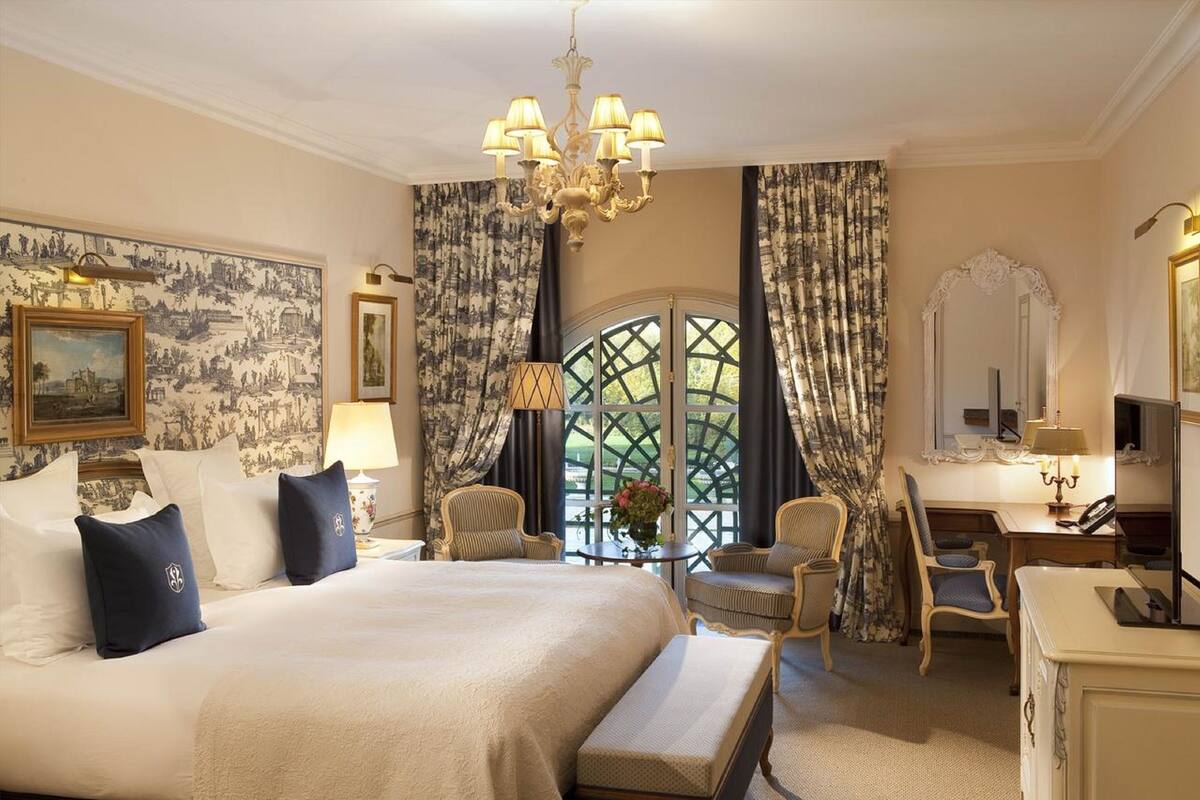 Ein Hotelzimmer mit einem großen Bett, zwei Stühlen, einem Schreibtisch, einer Kandelaber und einem Fenster mit Vorhängen.
