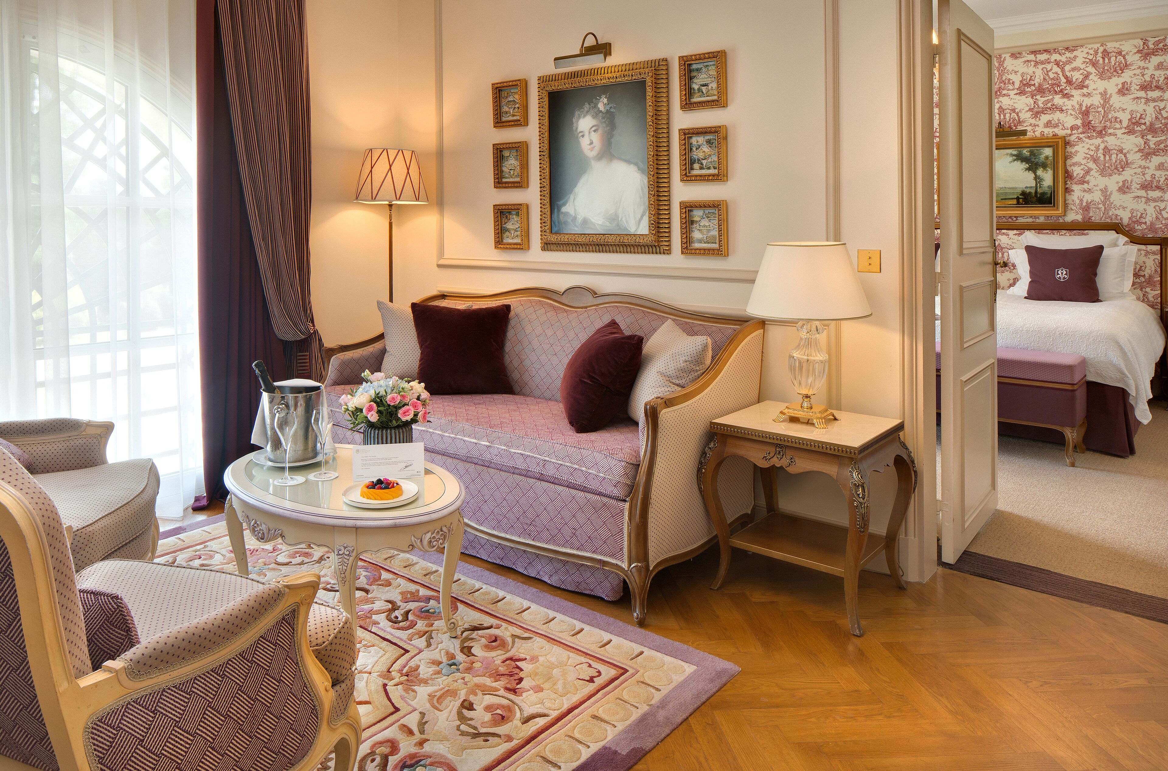 A room with a sofa, armchair, small table with a pitcher and flowers, and a bed with a nightstand.