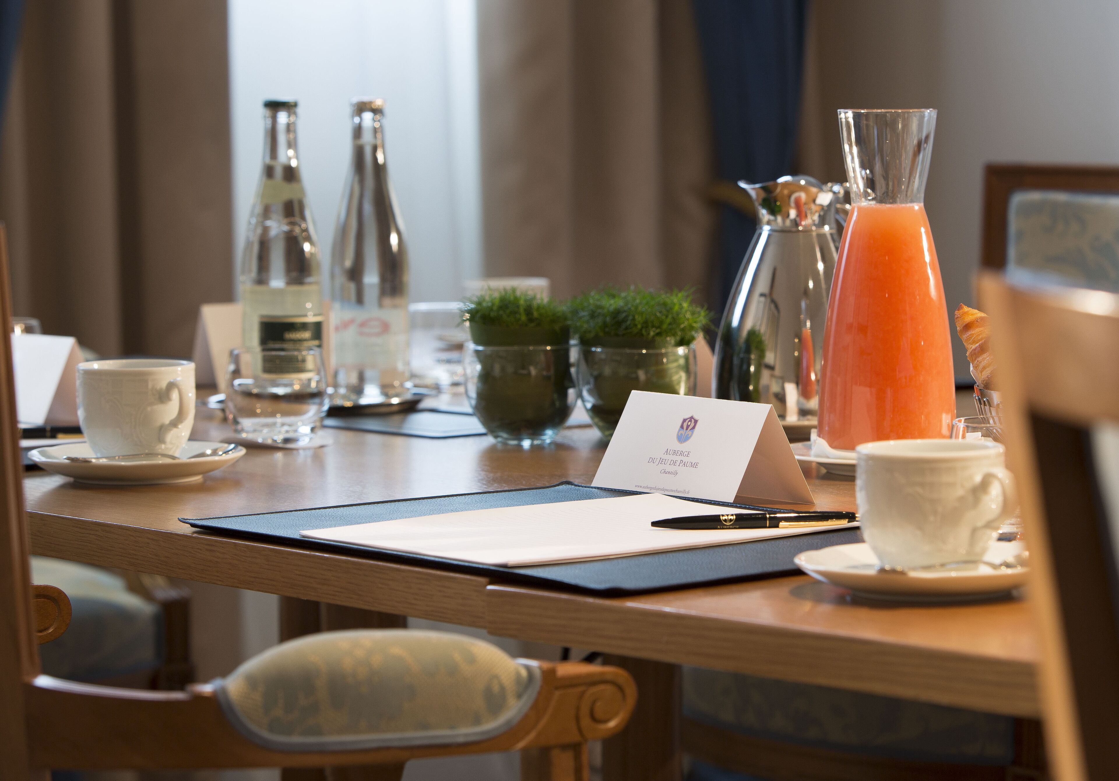 A hotel room with a wooden desk, a white cup, a glass of orange juice, a bottle of water, a small potted plant, and a business card.