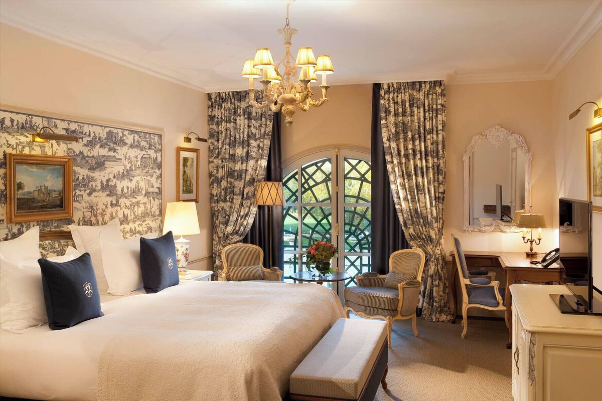 Ein Hotelzimmer mit einem großen Bett, einem Schreibtisch, einem Sessel, einer Kandelaber und einem Fenster mit Vorhängen.