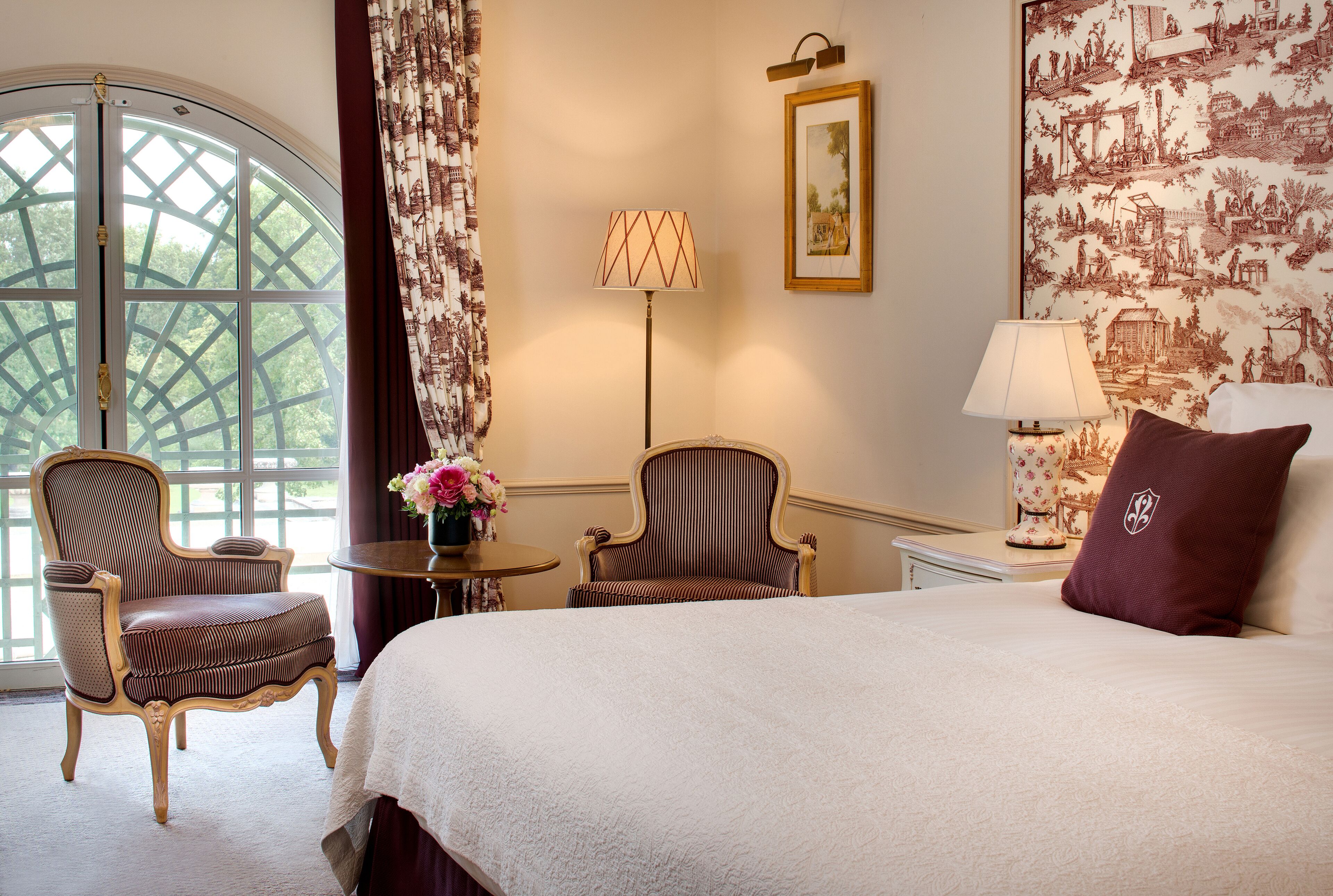 Una habitación de hotel con una cama grande, dos sillones, una mesita con un jarrón de flores, una lámpara y un amplio ventanal con cortinas.