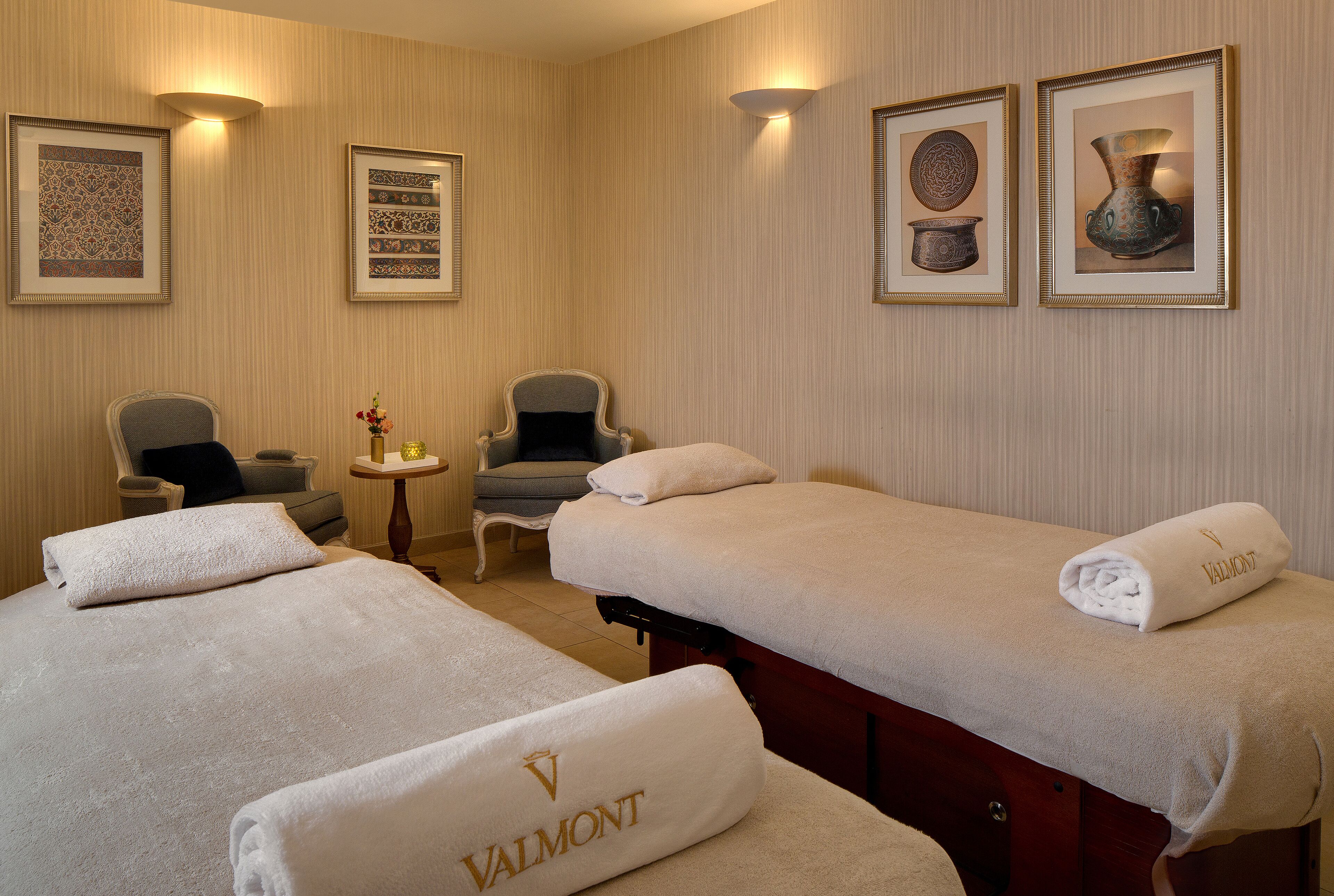 A hotel room with two beds, a small table, two armchairs, and wall-mounted artwork.