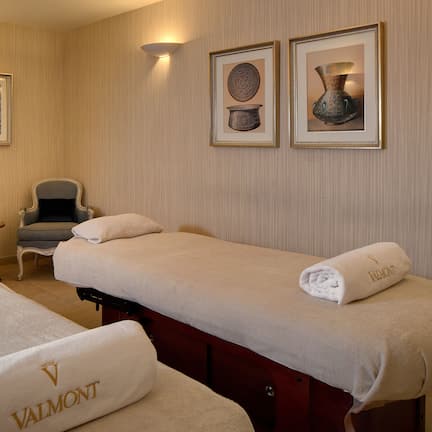 A hotel room with two beds, a small table, two armchairs, and wall-mounted artwork.