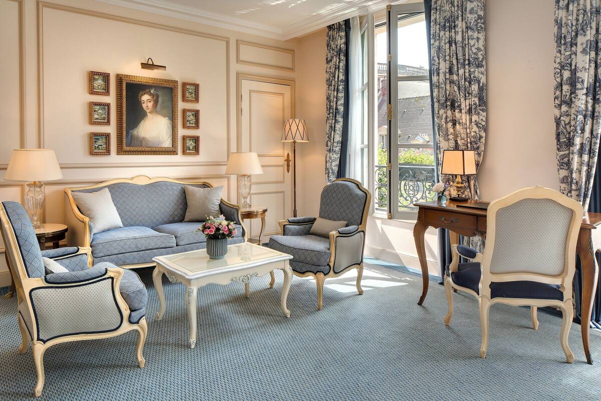Ein Zimmer mit einer Couch, Sesseln, einem Tisch und einem Schreibtisch. An den Wänden hängen gerahmte Bilder, auf dem Schreibtisch steht eine Lampe und der Blick fällt durch das Fenster.