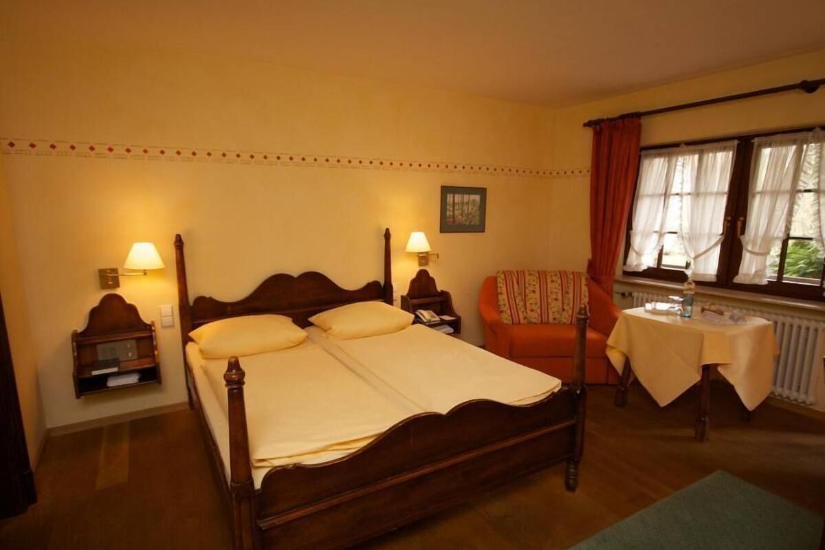 A bedroom with a wooden four-poster bed, two bedside lamps, a small table with a tablecloth, a chair, and a window with curtains.