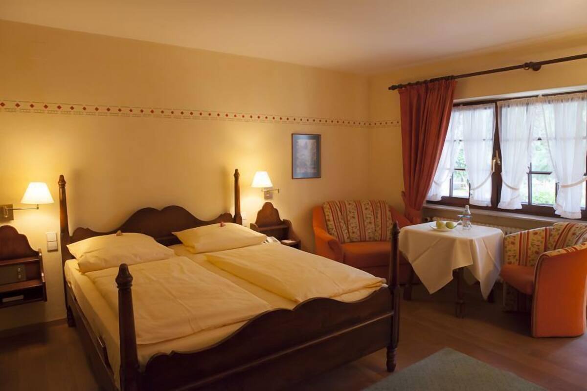 A hotel room with a double bed, two bedside tables with lamps, an orange armchair, a small table with a white tablecloth, and a window with curtains.