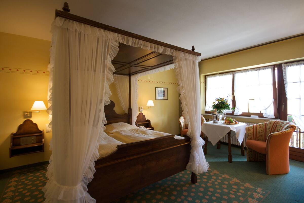 A four-poster canopy bed with a wooden frame and white drapes, a small bedside table with a lamp, a chair with a floral cushion, and a table with a vase of flowers.