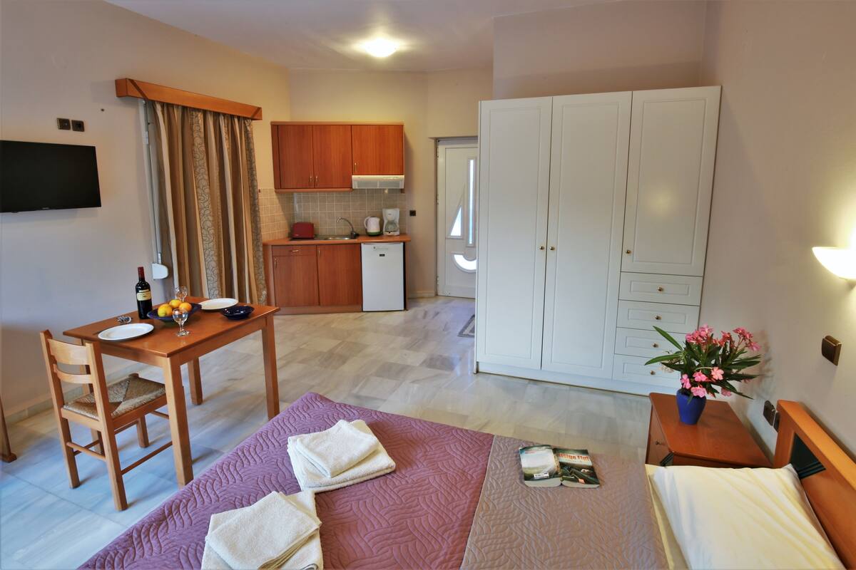 A compact hotel room with a bed, a small desk with a chair, a kitchenette, and a wardrobe.