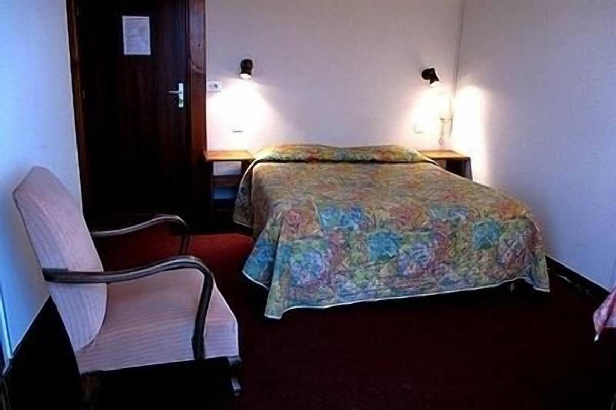 A small, dimly lit room with a single bed, a chair, and two wall-mounted lamps.