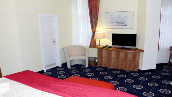 A hotel room with a bed, a desk with a television, a chair, and a patterned carpet.