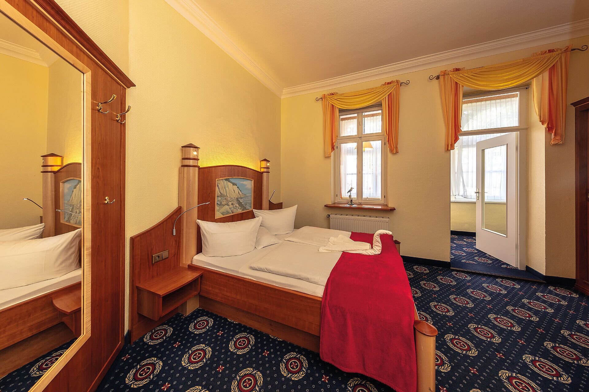 A hotel room with a wooden bed, a red towel, a painting on the headboard, and a window with curtains.