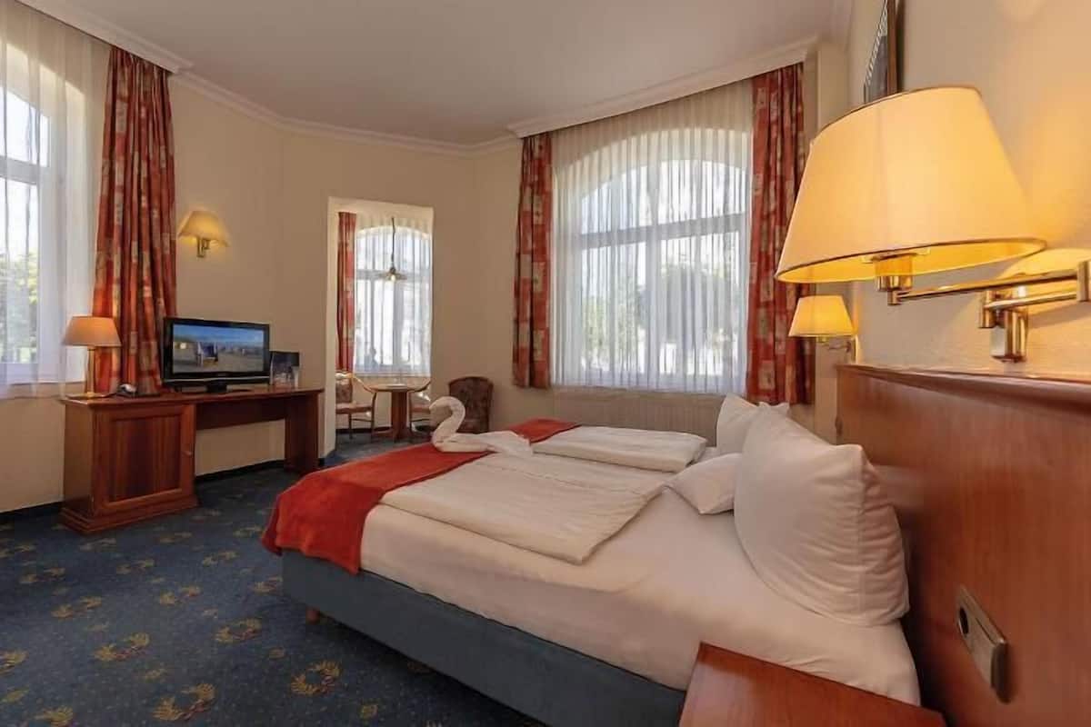 Habitación de hotel con una cama, un escritorio con televisor y dos ventanas con cortinas.