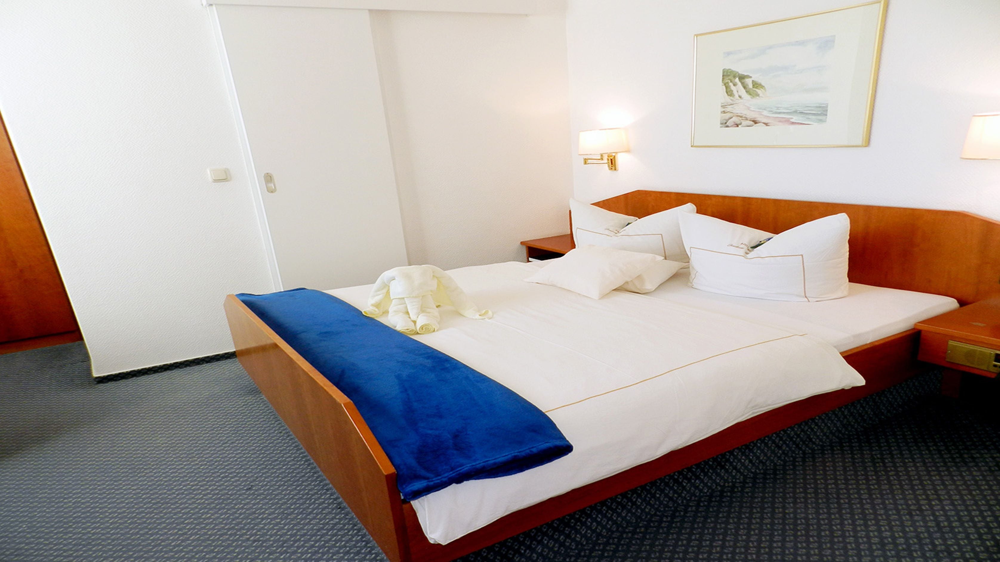 Habitación de hotel con cama de madera, ropa de cama blanca, una manta azul y un cuadro en la pared.