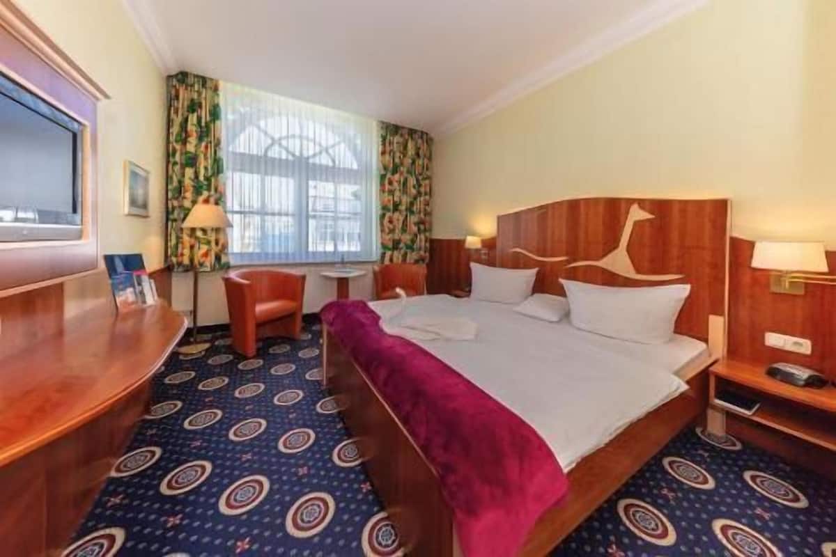 Una habitación de hotel con cama, escritorio, silla, una mesita y ventana con cortinas.