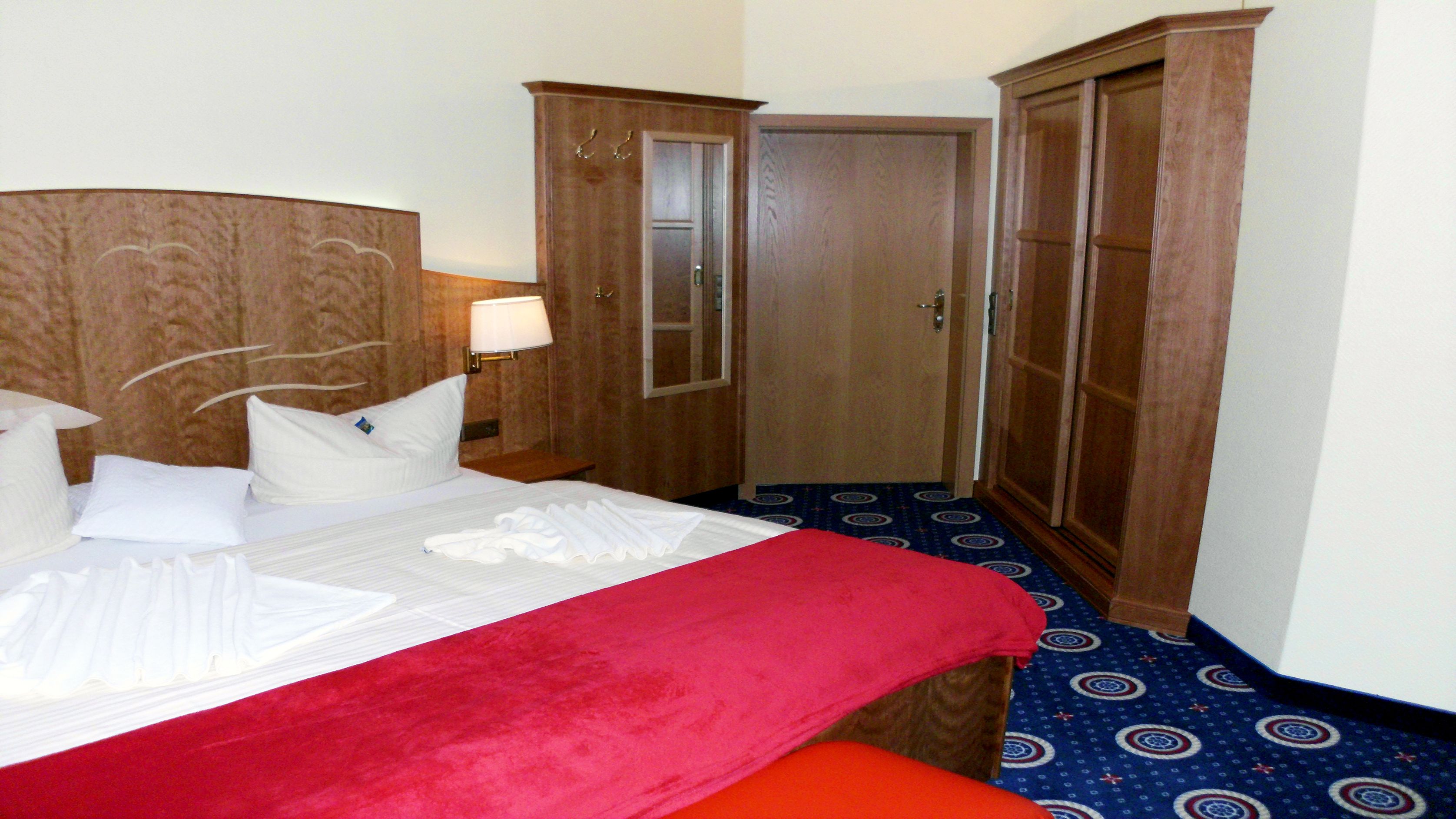 A hotel room with a bed, a red throw blanket, a wooden wardrobe, and a bathroom door.