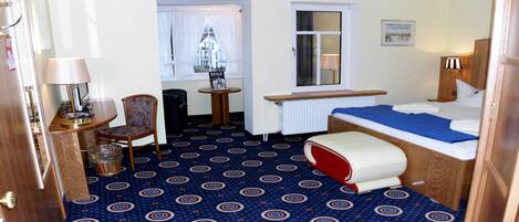 Habitación de hotel con cama, una silla, una mesita, una lámpara y ventana con cortinas.