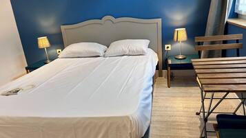Un lit bien fait, avec du linge de lit blanc, deux lampes de chevet, une chaise en bois et un mur bleu.
