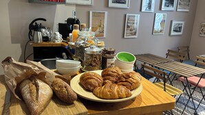 Desayuno con pan, croissants y recipientes con mermeladas sobre una mesa de madera, con libros y cuadros en la pared.