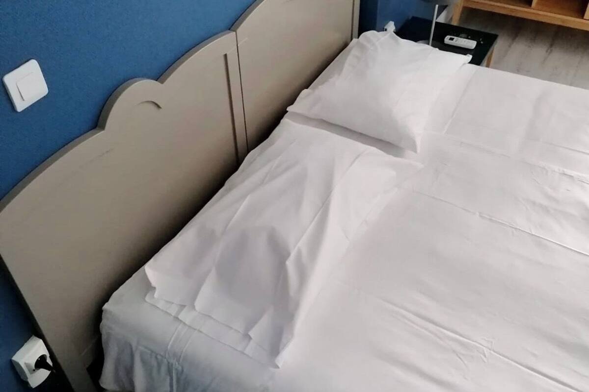Un lit avec une literie blanche, une tête de lit, une table de chevet avec une lampe et une prise électrique au mur.