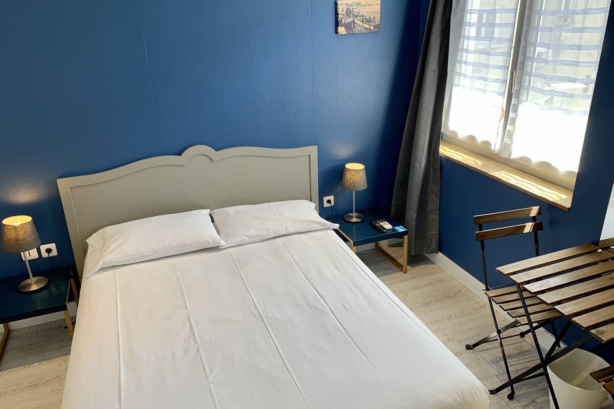Un lit bien fait, avec du linge de lit blanc, dans une pièce aux murs bleus, une petite table de chevet avec une lampe et un banc en bois.