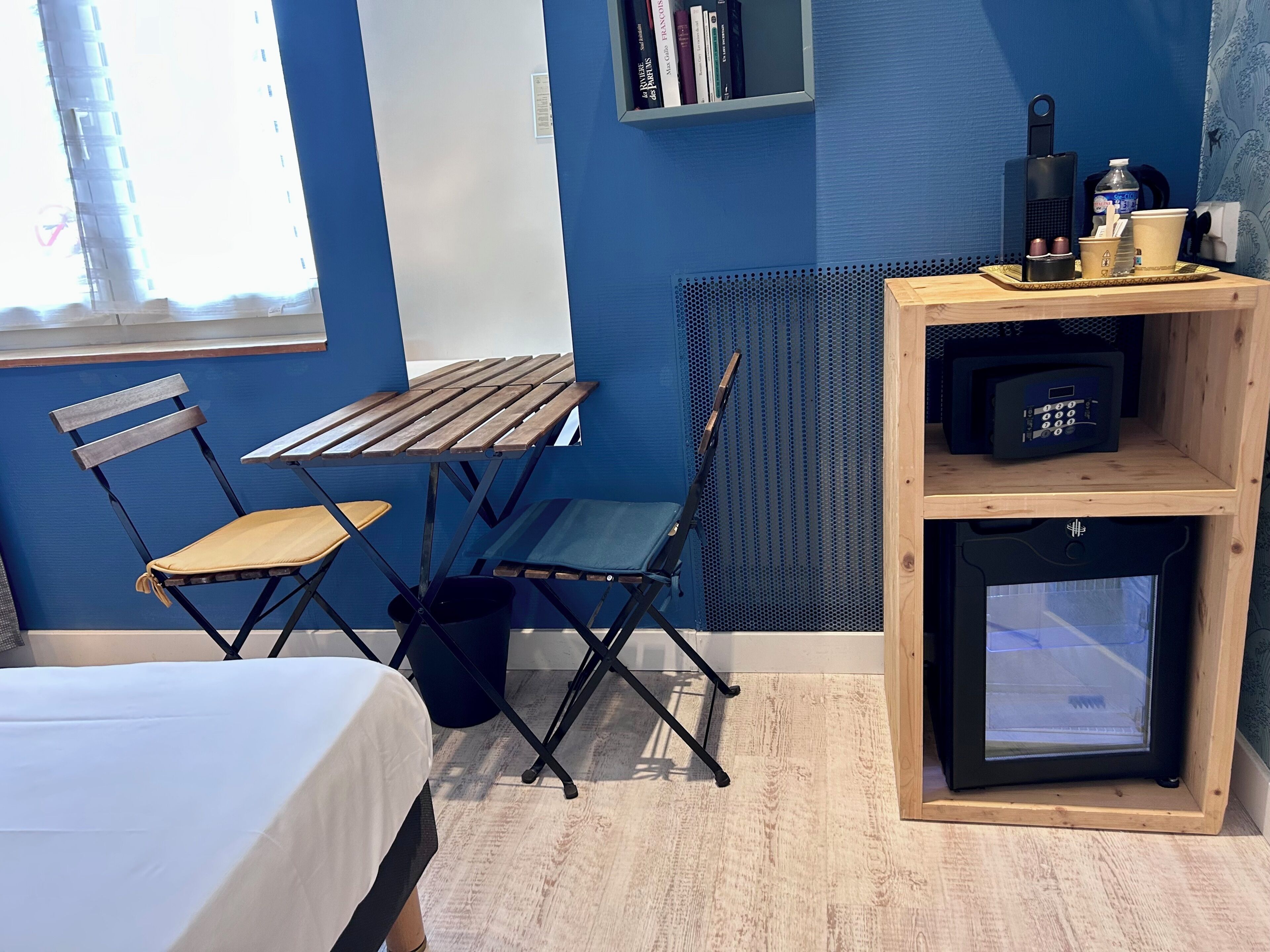 A compact room with a bed, a foldable table, two chairs, a radiator, a bookshelf, and a small refrigerator.