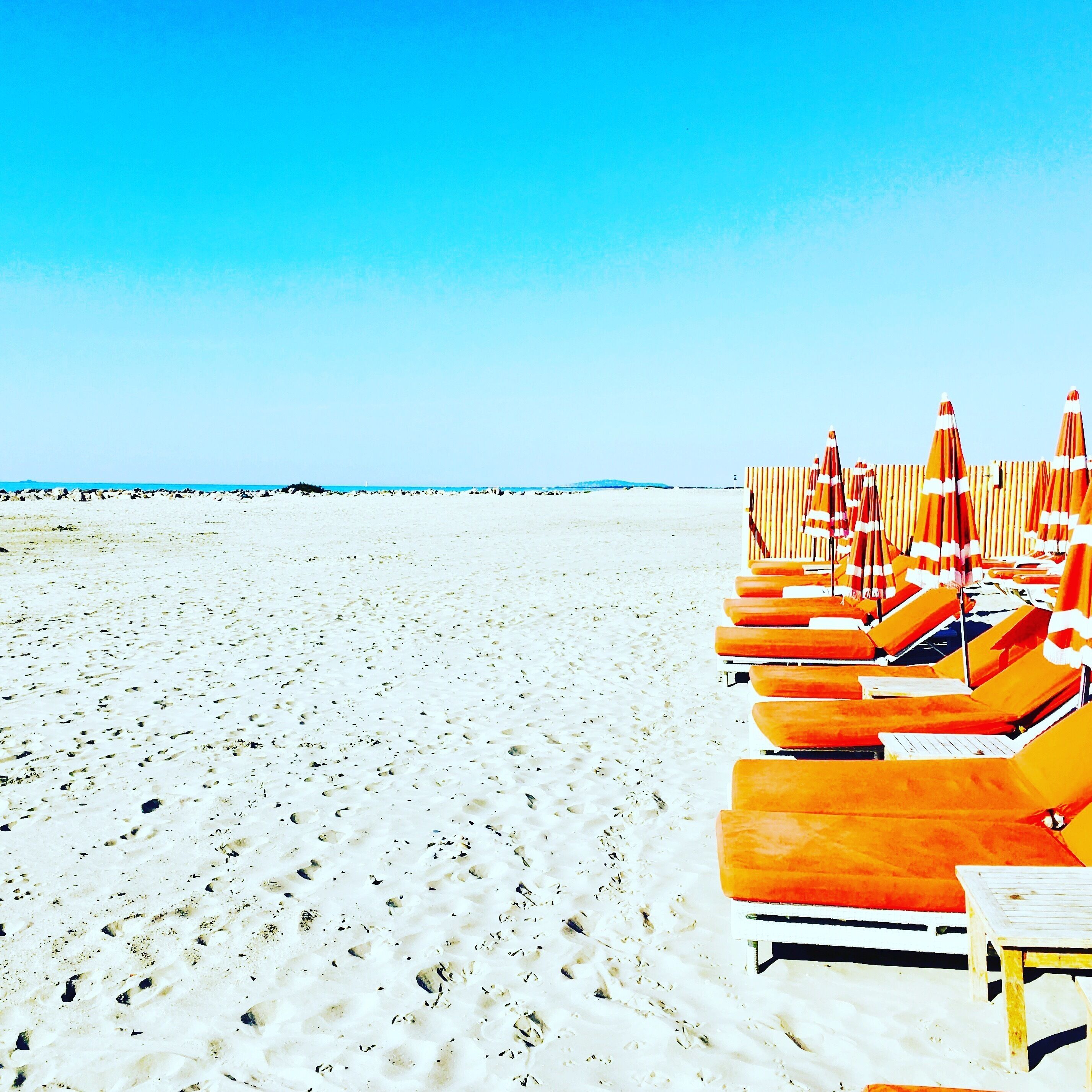 A beach with rows of orange sun loungers and umbrellas, a wooden table, and a clear blue sky.