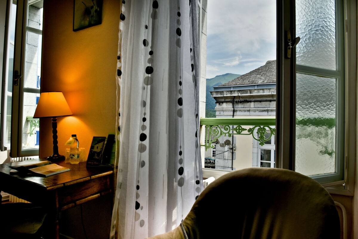 A room with a desk, lamp, and a view of a building through a window with a curtain.
