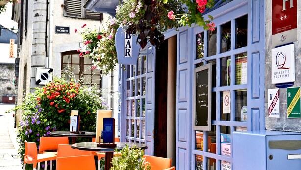 Un bar con tavoli all'aperto, un cartello con la scritta 'BAR' e diversi avvisi e cartelli sulla facciata dell'edificio.