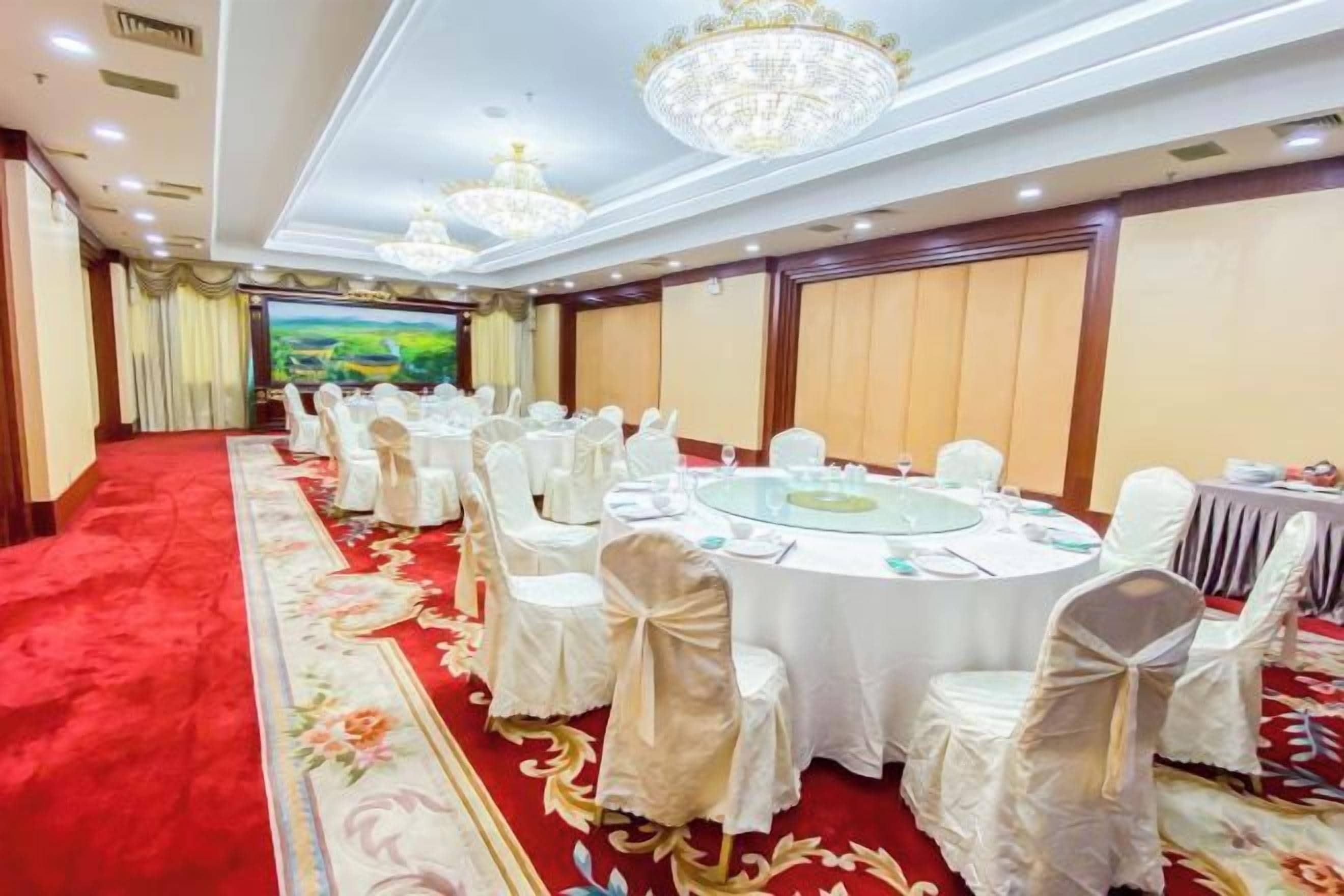Un salón de banquetes con mesas redondas, sillas blancas y alfombra roja.