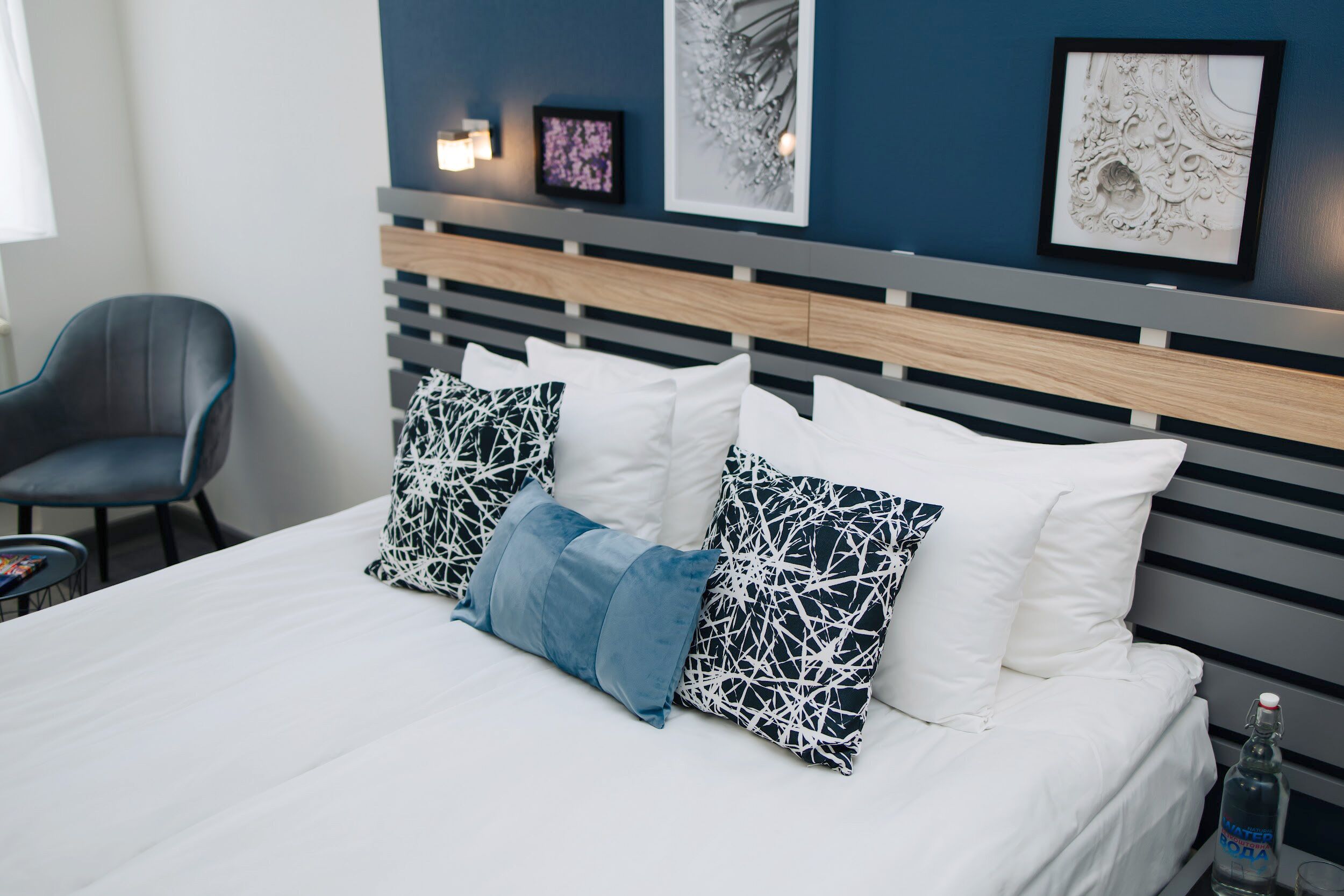A neatly made bed with white and patterned pillows, a grey upholstered chair, and wall art.
