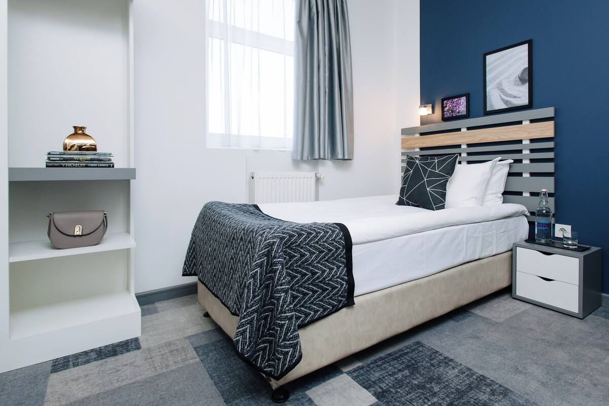 Un dormitorio moderno con cama, mesitas de noche, un radiador, una ventana con cortinas y una repisa en la pared con objetos decorativos.