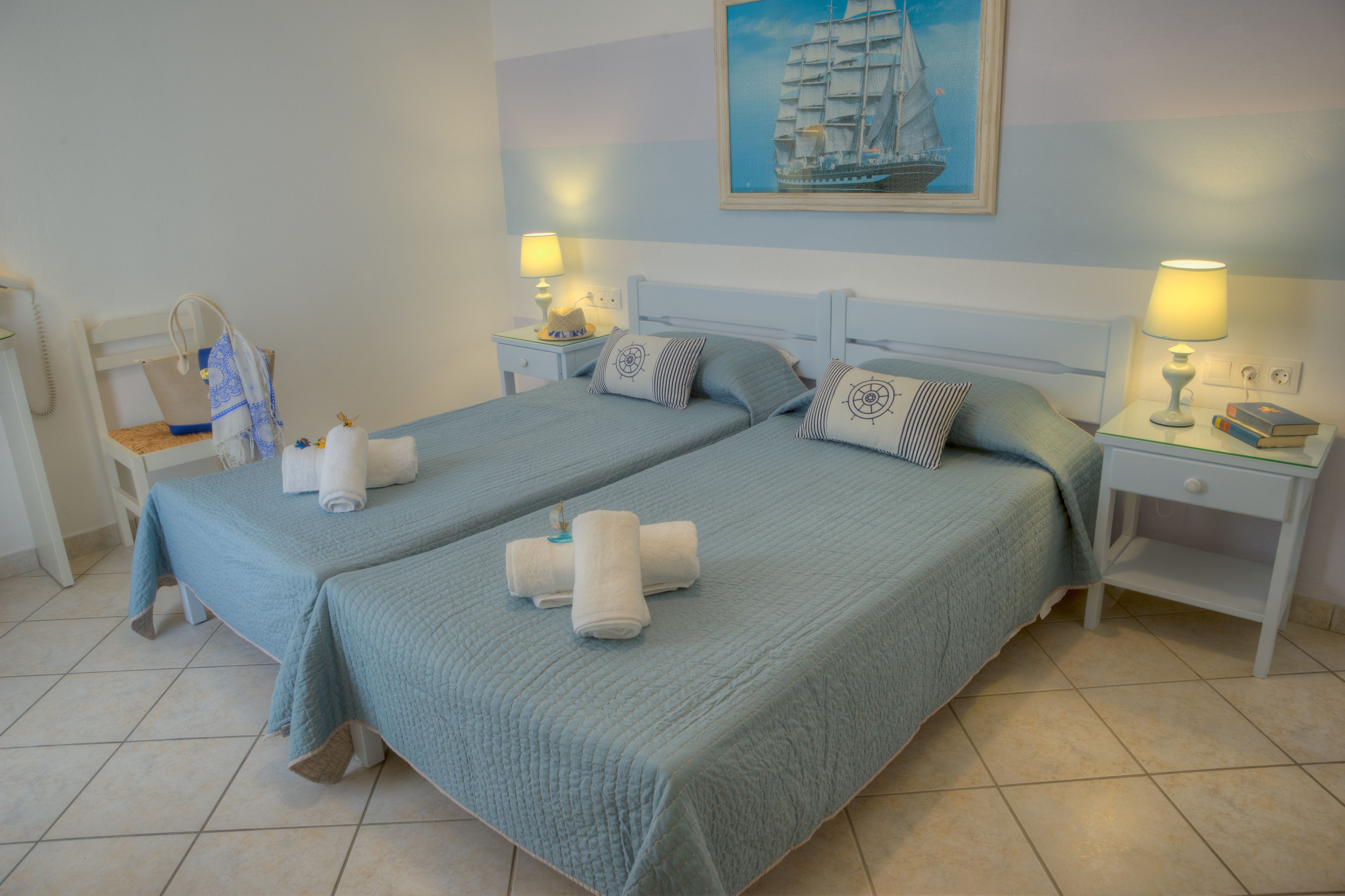 A hotel room with two beds, each with a blue bedspread and pillows, a painting of a sailboat on the wall, and bedside lamps.