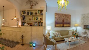 A well-lit living room with a sofa, coffee table, and chairs. There is a bookshelf filled with books and decorative items. - Cyclades Hotel (Paros)