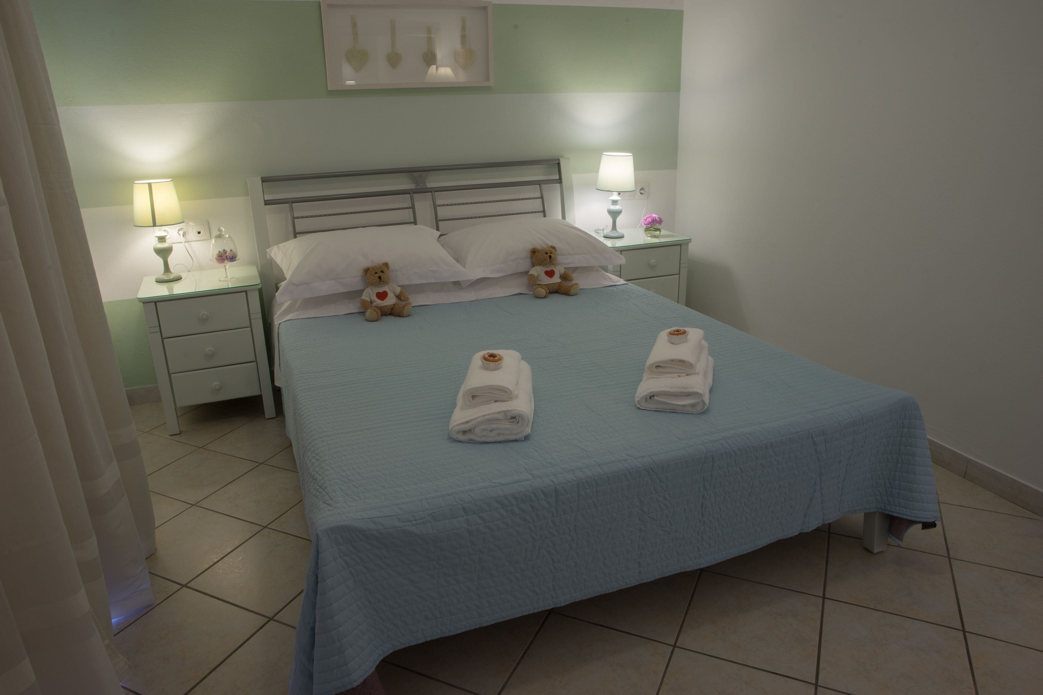 A neatly made bed with two towels, two bedside lamps, and two teddy bears on the pillows.