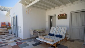 A sunlit room with a striped sofa, a small table, and a chair. A door and a wall-mounted shelf with items are also visible. - Cyclades Hotel (Paros)