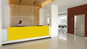 A reception desk with a wooden panel wall, a receptionist, and a bright yellow counter. - Amaris Hotel Juanda (Jakarta)