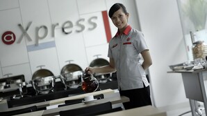 A person in a uniform pouring tea into a cup in a modern kitchen with stainless steel appliances and a branded wall. - Amaris Hotel Juanda (Jakarta)