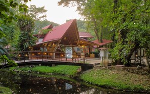 A restaurant with a red roof and a signboard, surrounded by lush greenery and a small bridge over a pond.