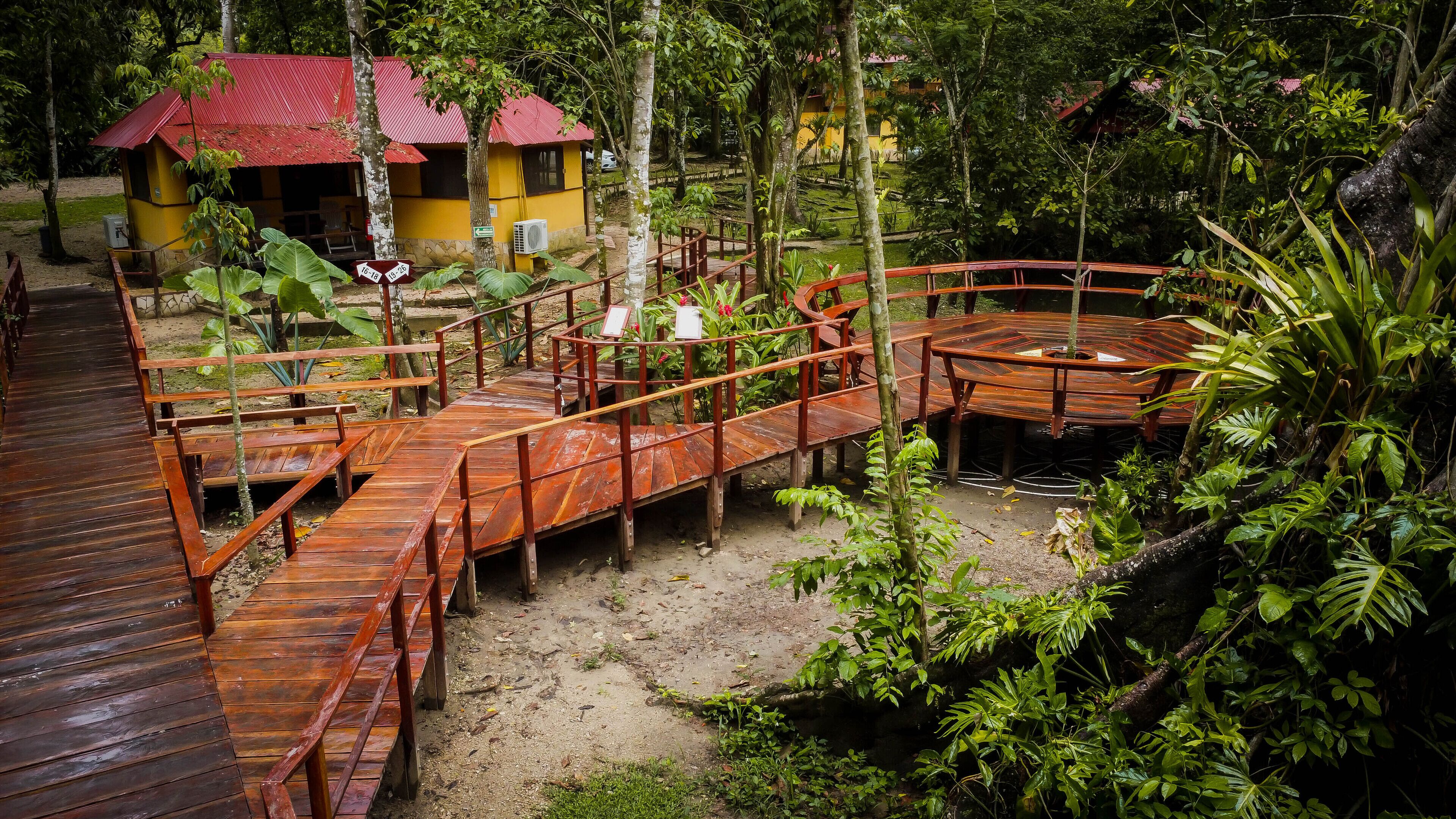 Una terraza de madera con un área de estar circular, rodeada de abundante vegetación y un pequeño edificio con techo rojo.