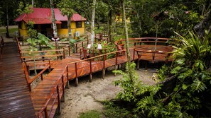 A wooden deck with a circular seating area, surrounded by lush greenery and a small building with a red roof.